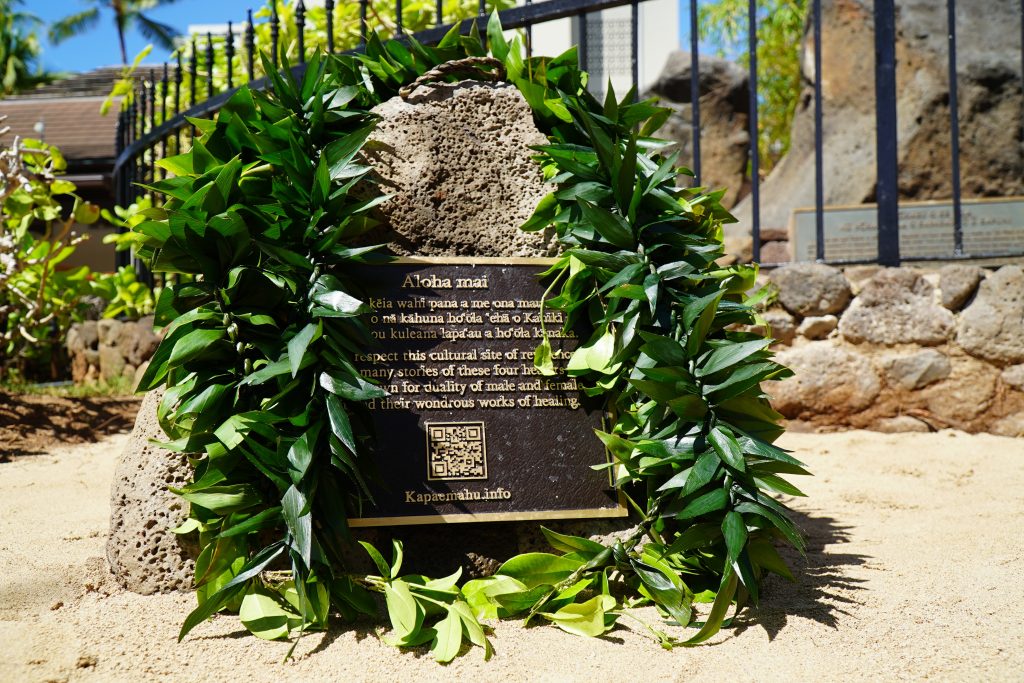 A stone with a plaque is draped with lei outside the Kapaemahu Stones in Waikīkī. 